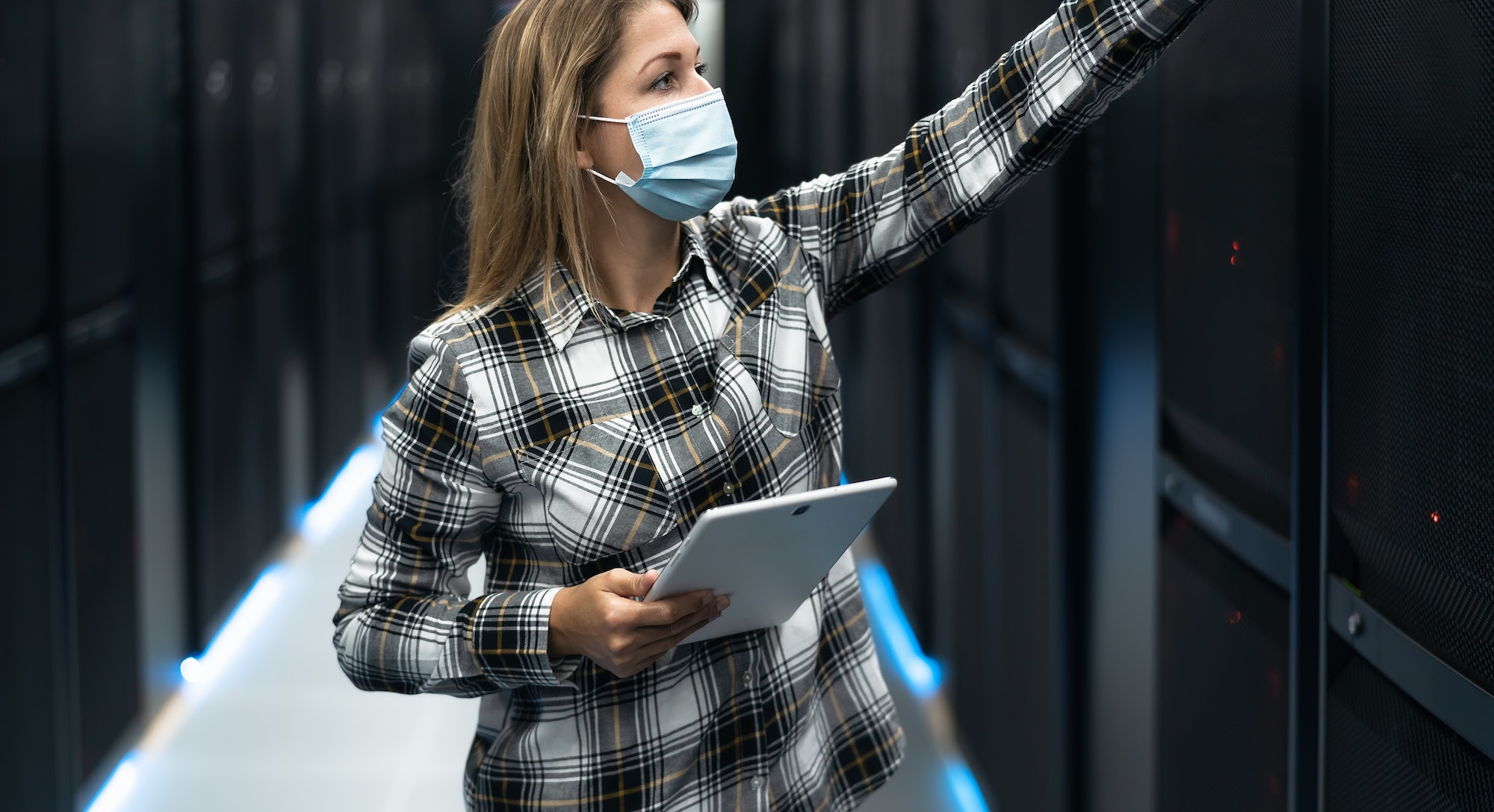 Female data center technician working inside server rack room while wearing face mask