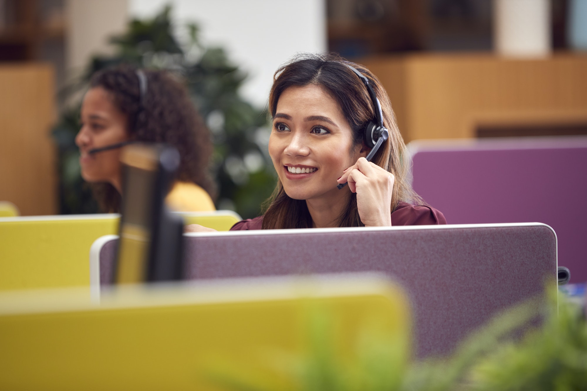 Mature Businesswoman Wearing Phone Headset Talking To Caller In Busy Customer Services Centre