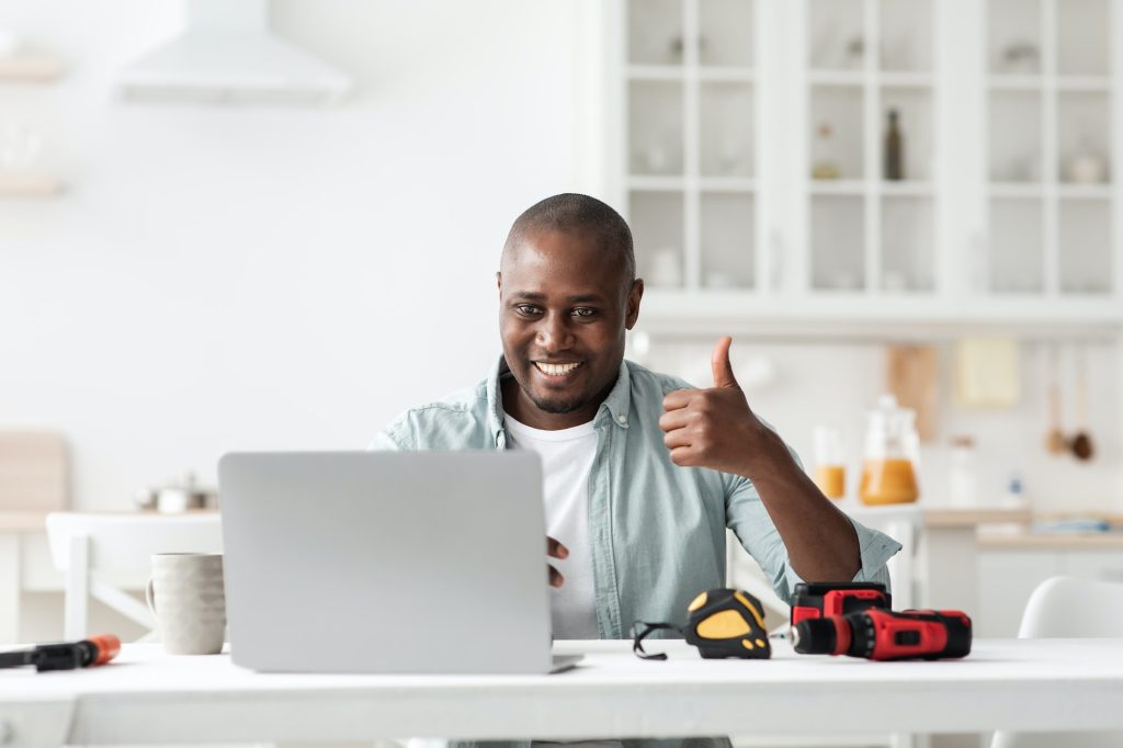 Review of construction instruments. Happy black handyman looking at laptop webcam and showing thumb